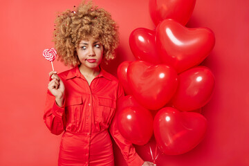 Obraz na płótnie Canvas Holiday concept. Sad dissatisfied young woman with curly hair wears dress holds lollipop and bunch of inflated heart balloons isolated over vivid red background feels very tired after party.