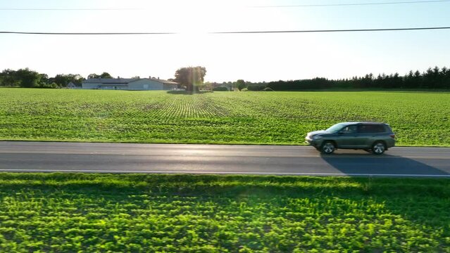 Tracking shot of SUV traveling along rural road. Fields surround car as sunlight reflects off its windshield. Travelers cross farmland at sunset.