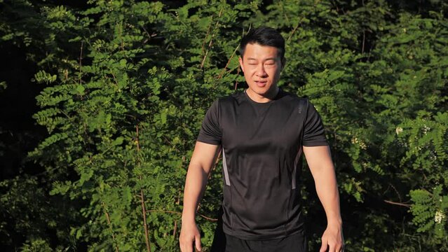 Front view of active fit young korean man doing squat exercise during workout at street sunset background of green trees. Summer time male workout sport outdoor. Sport healthy concept.
