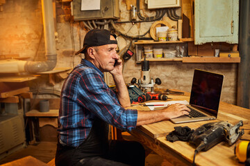 Senior carpenter works on the computer and talks phone with client in the workshop