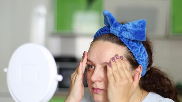 Curly woman in hairband applying and smearing moisturizing lotion or skincare cream for skin lifting. Looking in mirror, face self massage. Circle and patting movements, home spa procedures treatment