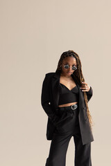 African girl in a black suit with long hair and sunglasses stands on a beige background