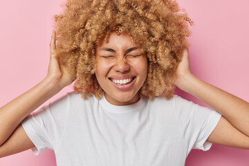 Studio shot of cheerful young woman keeps hands on her natural curly hair closes eyes and grins at camera expresses positive emotions wears casual white t shirt isolated over pink background