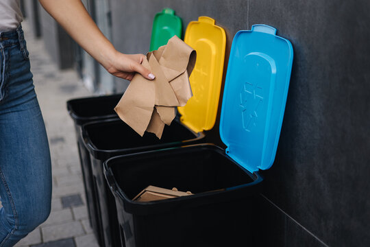 Close-up of female throwing piece of cardboard in recycling bin. Different colour of recycling bins outdoors. Side view of woman put rubbish in container