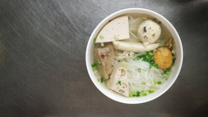 Pho, Vietnam rice noodles with pork ribs soup, vietnamese sausage and fish ball serve in white plastic bowl. On stainless table at street stalls. Top view and copy space.