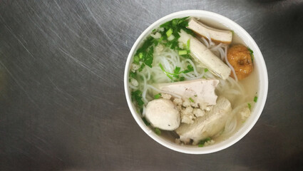 Pho, Vietnam rice noodles with pork ribs soup, vietnamese sausage and fish ball serve in white plastic bowl. On stainless table at street stalls. Top view and copy space.