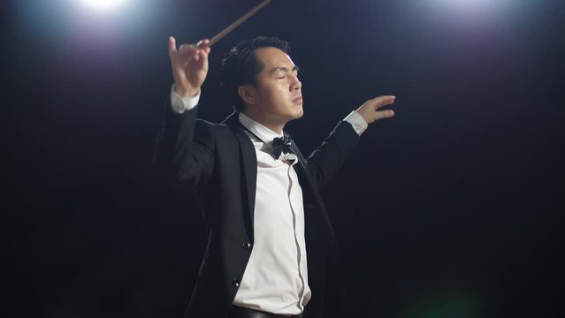 Side View Of Asian Conductor Man Holding A Baton Closing His Eyes And Showing Gesture In The Black Studio
