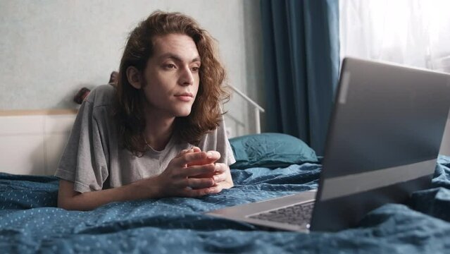 Young man lies on his stomach in bed and looks at the laptop screen. His face is relaxed, his eyes are attentively looking at the screen. Slow motion 4k footage