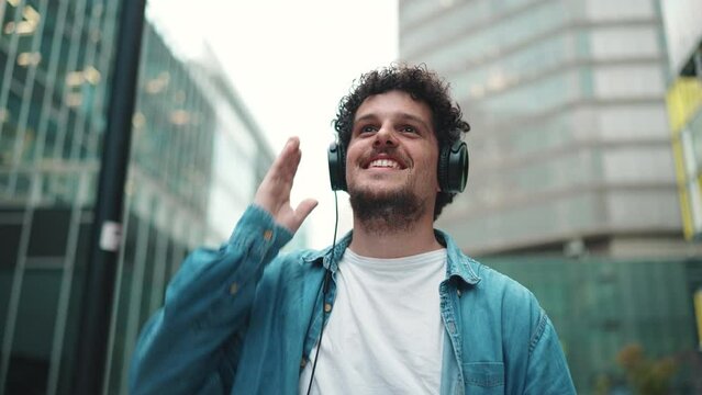 Close-up of young bearded man in denim shirt walking down the street wearing headphones listening to music on modern cityscape background
