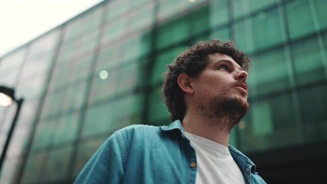 Close-up, man in denim shirt walks down the street, stops and looks around at the upper floors of modern buildings