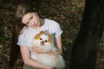Young woman with long straight hair and fluffy small dog for walk in forest. Pomeranian with his owner for walk. Satisfied dog smiles and sticks out his tongue. Woman and dog looks at camera