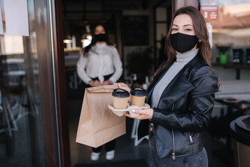 Attractive female customer in face mask standing outdoors by cafe and take away food and coffee. Background of cashier in mask waiting for next customer. New norm during quarantine