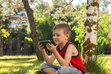 little boy uses smartphone while sitting on green grass in the park. modern childhood concept