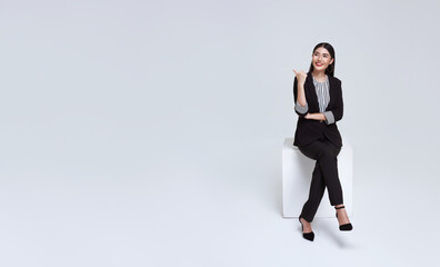 friendly face asian businesswoman smile in formal suit sitting on chair and points her hands presented to copy space on white background studio shot.