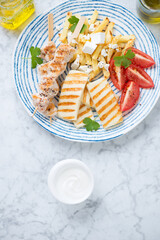 Plate with grilled halloumi, souvlaki, greek-style fries with feta and tomatoes, above view on a light-grey marble background, vertical shot