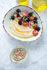 Bowl of hummus with tomato salad on a light-grey marble background, elevated view, vertical shot