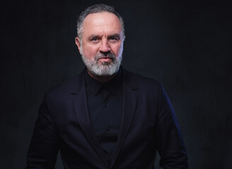 Studio shot of stylish aged man with beard dressed in black coat and black shirt.