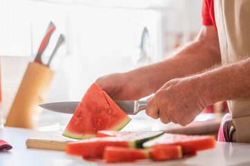 Caucasian senior man in home kitchen while cutting a ripe red watermelon in slices - active retiree dressed in red, summer fruit concept - Powered by Adobe