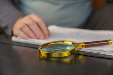 A man's hand is leafing through documents. A round magnifying glass with a gold-colored frame lies in the foreground. A clipboard for papers. Inside the room. Selective focus.