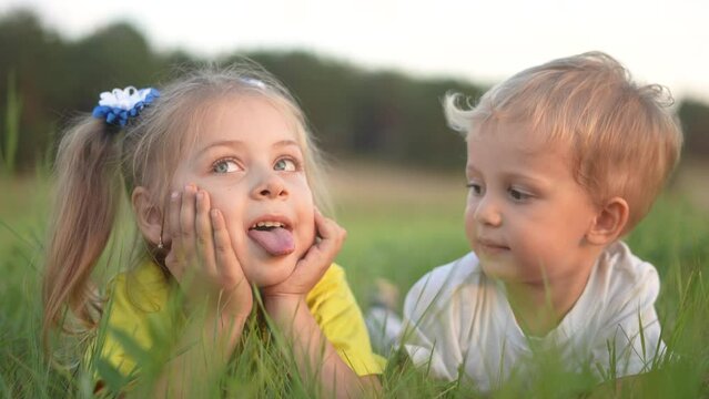 little girl showing her tongue to her brother lying on the grass in the park. happy family kid dream concept. fun small children play in the summer in nature in the park in the green grass