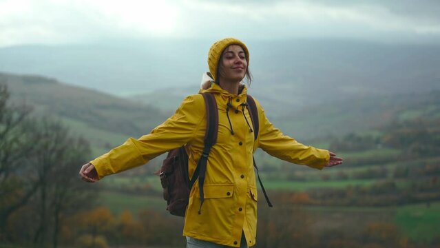 Beautiful woman in yellow raincoat and backpack stands against fields and mountains, raises her arms, inhale and breathe in fresh after rain air. Travel adventure Scandinavian tourism concept.