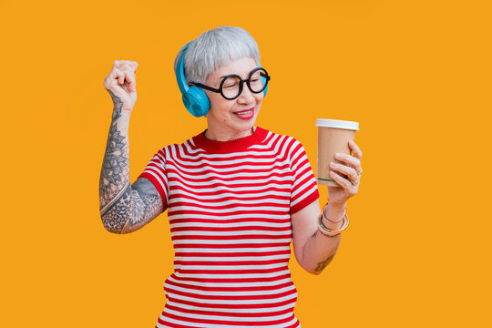 Cheerful elderly gray-haired asian female woman with tattoo casual cloth listening music with headphones keeping eyes closed isolated on pastel yellow color background studio portrait