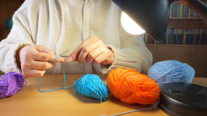 Close up of female hands holding knitting needles. Woman knitting homemade things in dark room under lamp. Diy hobbies.
