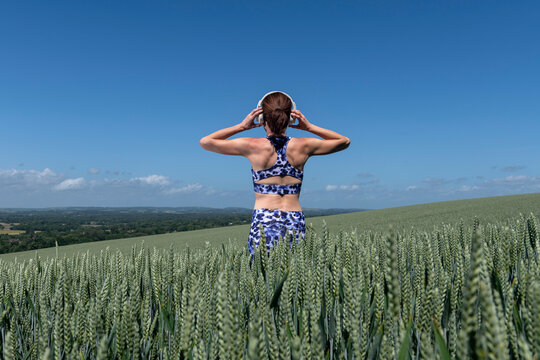 Sporty, fit woman listening to music with wireless headphones in a field. Rear view