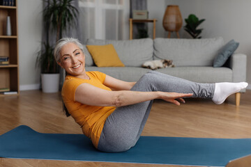 Happy fit senior woman doing abs exercises on yoga mat at home, working out her core muscles, doing...