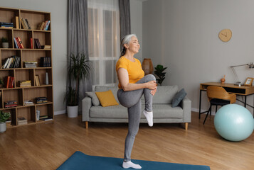 Domestic fitness. Fit senior woman in sportswear doing exercises on yoga mat, stretching her leg on...