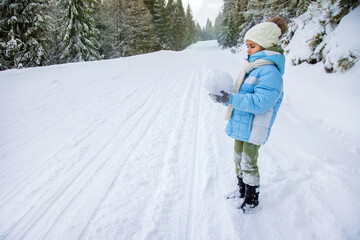 Fototapeta na wymiar A girl in a jacket and hat stands on the road in the forest and holds a snowball