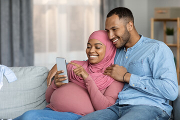 Happy Pregnant Black Islamic Couple Using Smartphone While Relaxing Together At Home