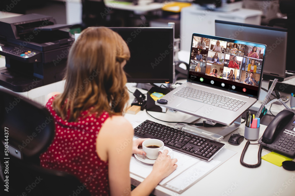 Canvas Prints Caucasian businesswoman holding coffee cup video conferencing with colleagues on laptop in office