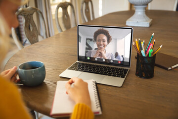 Caucasian businesswoman writing in notepad while video conferencing with colleague over laptop