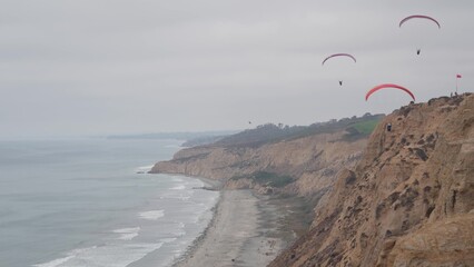 People paragliding, Torrey Pines cliff or bluff. Paraglider soaring in sky air on parachute, kite or wing. Sport hobby. Ocean coast, San Diego, California USA. Para gliding risky flight. Birds flying.