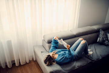 A woman lying on sofa and relaxing with interesting book at home.