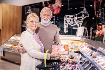 Happy senior couple showing stakes they buying at meat department in supermarket and smiling at the camera.