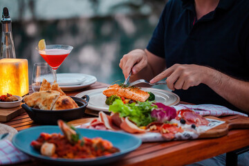 Man on luxury dinner served on table with fresh seafood, cold cuts and cocktail glass in luxury outdoor restaurant. Man hands cutting grilled salmon. Healthy food
