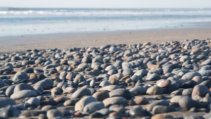 Fototapeta premium Pebble rocks on sandy beach by ocean sea water waves, round smooth stones or boulders low angle view in soft focus. California coast vacations aesthetic, USA. Seascape natural background with bokeh.