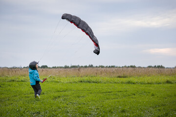 A cute kid in a blue jacket runs across a green field, launches a training parachute wing, holds...