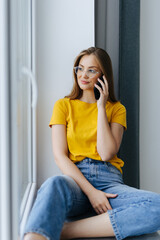 Young woman talking on the smartphone while sitting on the windowsill at home, chatting with friends.