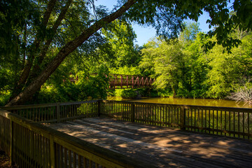 a brown wooden deck over the silky brown waters of Little River surrounded by lush green trees, grass and plants with a metal rust colored bridge and blue sky at Olde Rope Mill Park in Woodstock