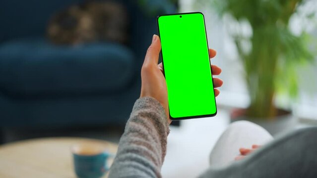 Woman at home using smartphone with green mock-up screen in vertical mode. Girl browsing Internet, watching content, videos. POV. Cat is lying on the chair on the background