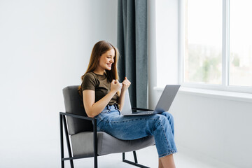 Overjoyed woman sitting in armchair with laptop, celebrating huge online win or success against white studio wall, copy space. Young lady enjoying big sale in web store