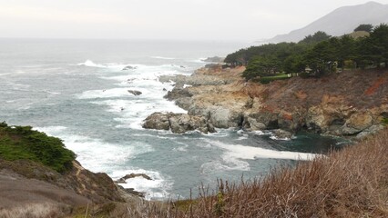 Fototapeta na wymiar Rocky craggy ocean shore, foggy misty weather. Sea water waves crashing on beach. California scenic landscape, Big Sur nature, USA. Pacific coast highway seascape viewpoint from cliff or steep bluff.