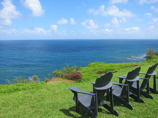 Relaxing concept. Three blue wooden chairs on a cliff's green lawn facing a deep blue ocean. Blue sky with white clouds. Princeville, Kauai, Hawaii.