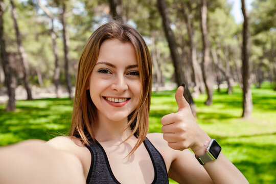 Happy sportive woman wearing sports bra standing on city park, outdoors showing thumb up and taking selfie photo. Looking at camera.