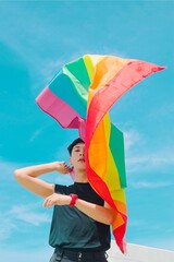 Young man  standing over beautiful blue sky and waving lgbti+ flag.