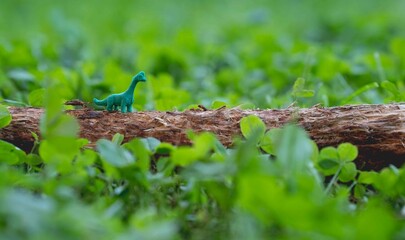 A green dinosaur toy stands on a tree trunk against a background of green grass