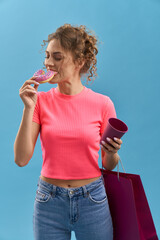 Curly woman eating tasty donut, while holding paper bag in studio. Portrait of pretty girl enjoying...